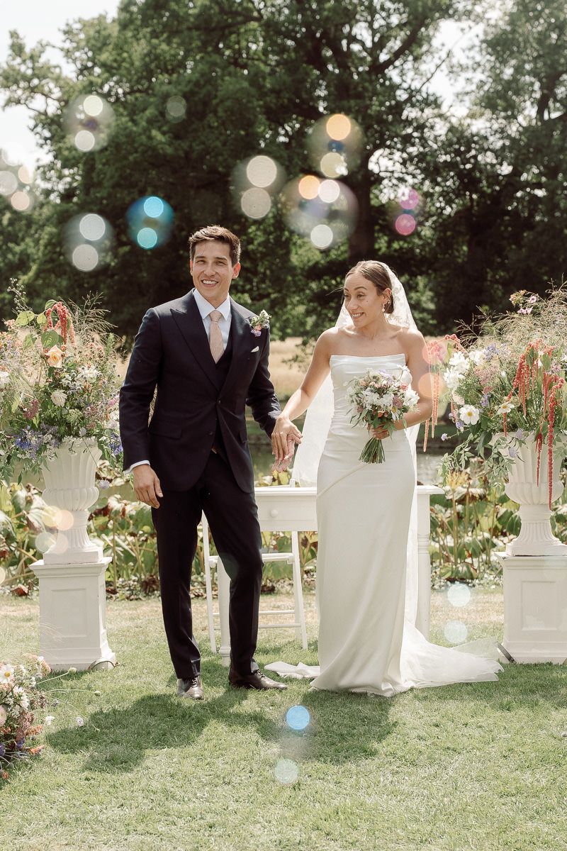 A romantic wedding couple poses playfully in a garden setting with white pillars and blooming flowers.