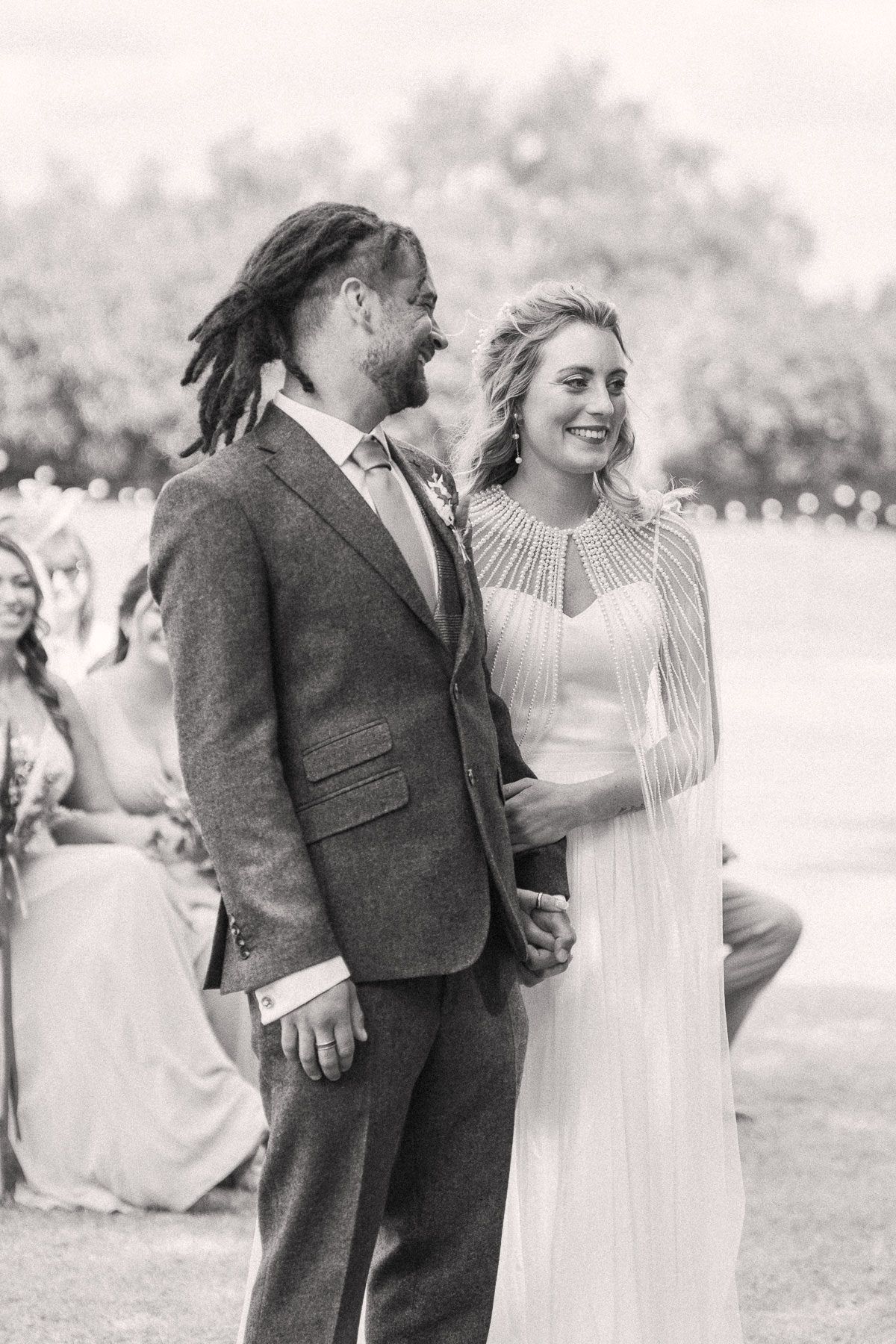 A black and white image showing a couple standing together, the woman wearing a long white dress and the man wearing a suit.
