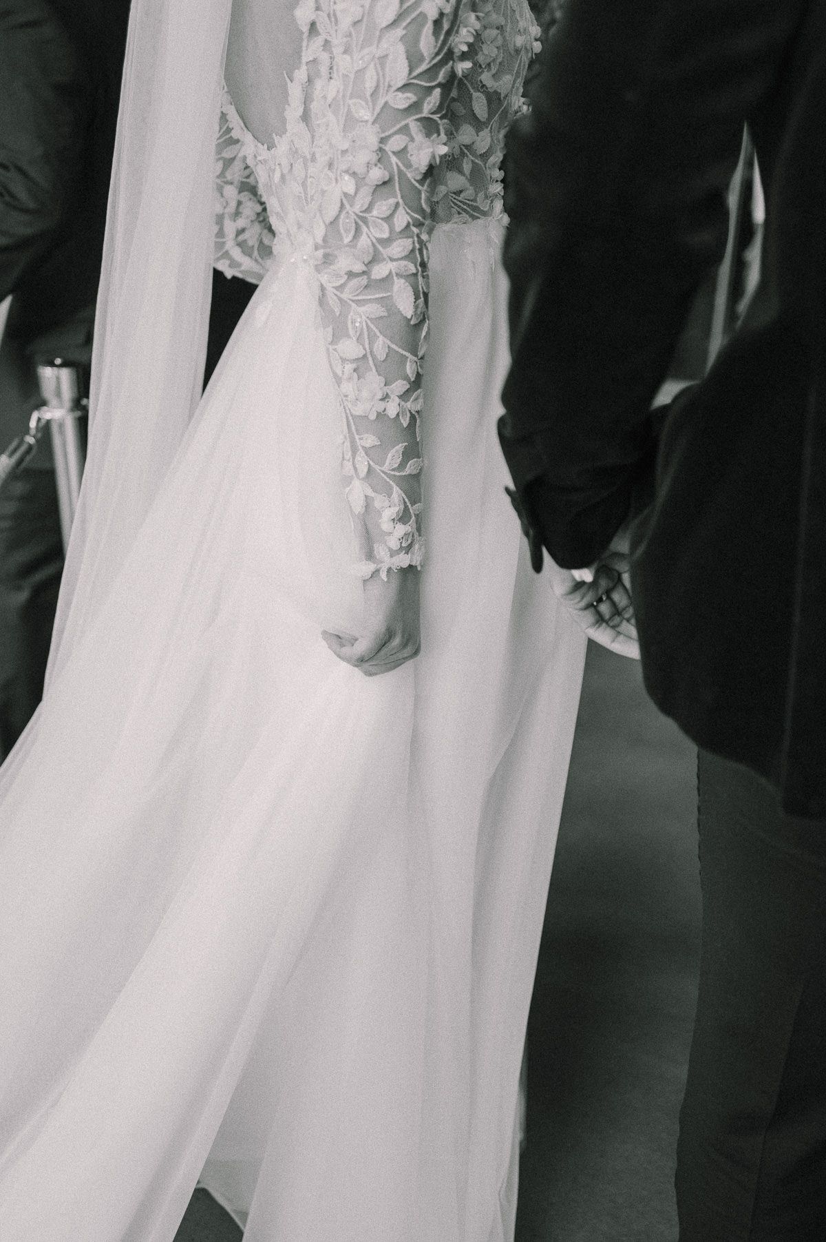 Black and white photo of bride and groom holding hands, showcasing intricate floral lace wedding gown with flowing veil.