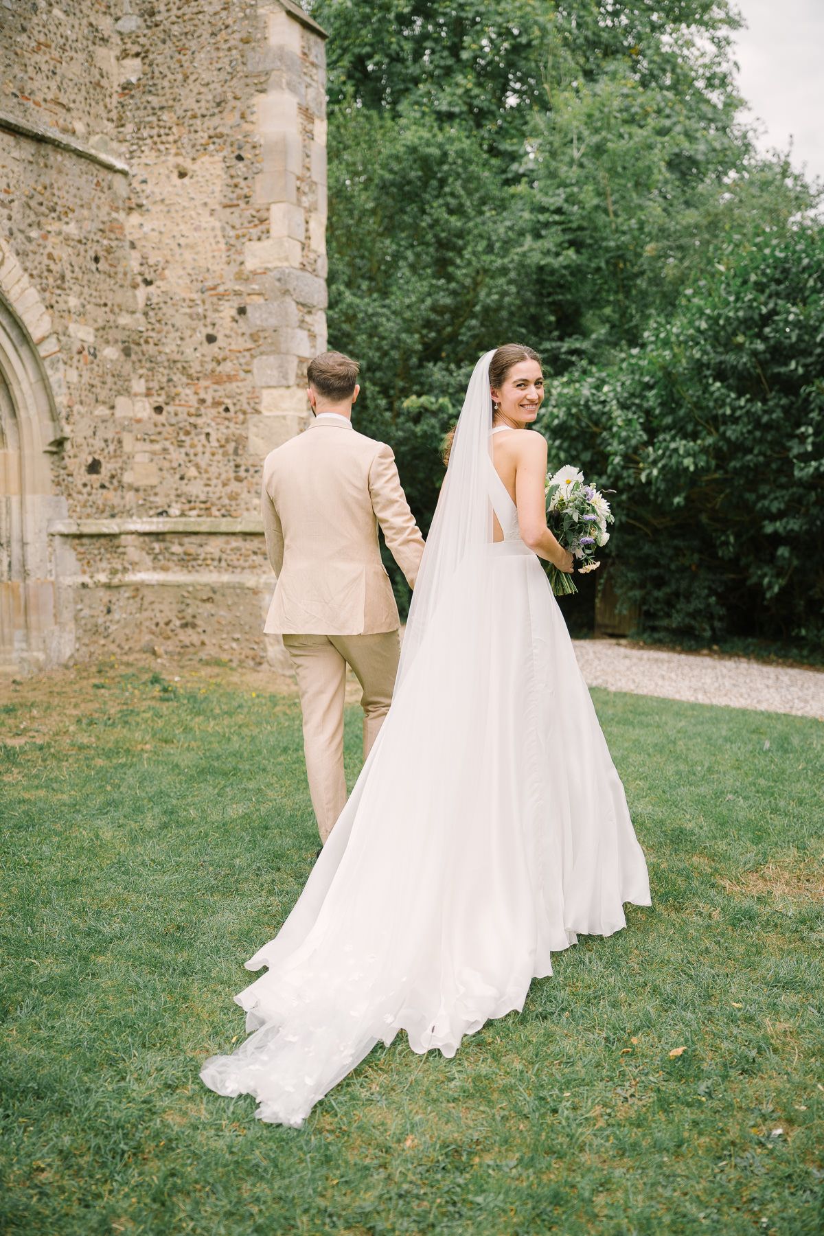 A bride and groom holding hands while walking on the grass in front of a stone building.
