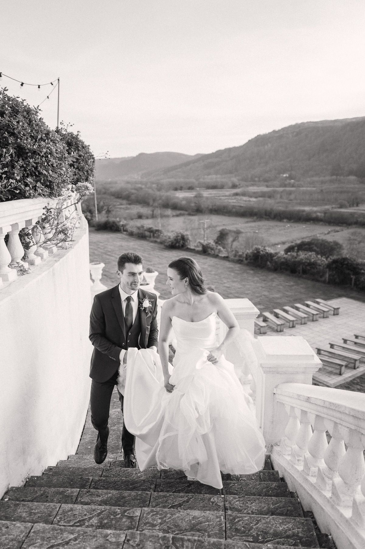 A bride and groom walk down outdoor stone steps overlooking a scenic landscape in this black and white wedding photograph.