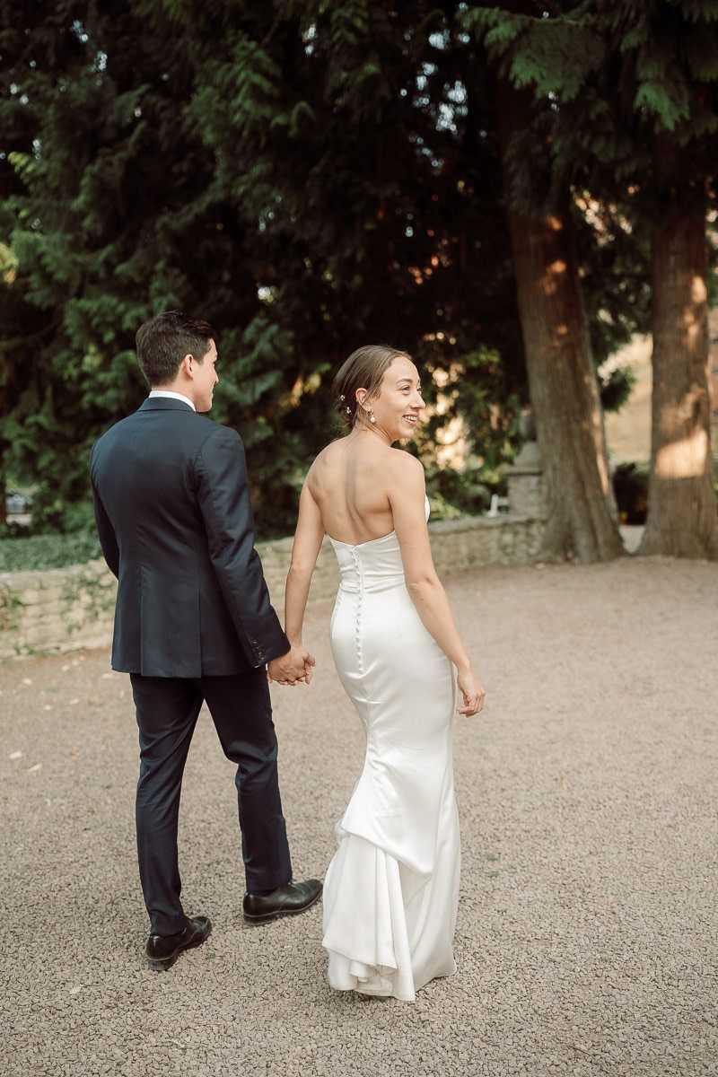 A couple in wedding attire walks together on a gravel path through a garden setting.