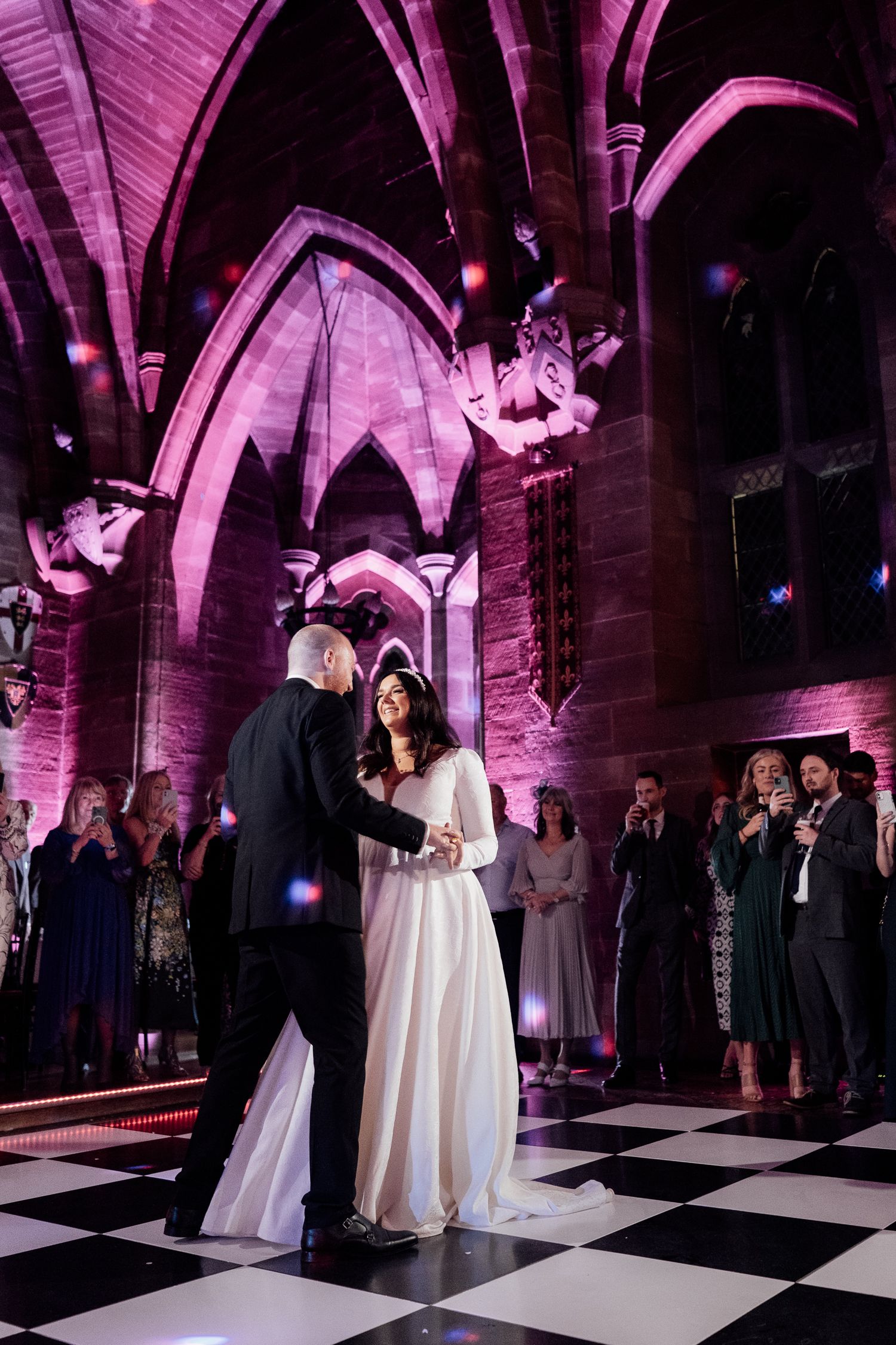 A couple shares a dance on a black and white checkered floor under dramatic purple lighting in a gothic-style venue.