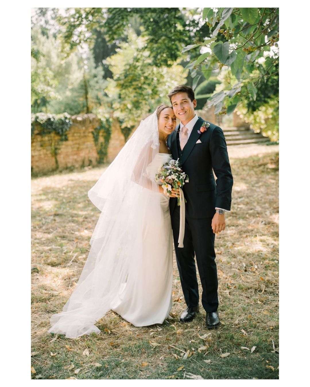 Bride and groom pose together outdoors in garden setting, bride wearing white gown with veil holding bouquet.