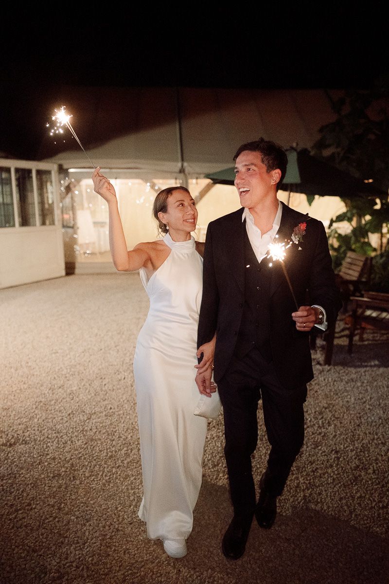 A couple in formal attire walks together at night holding sparklers during a wedding celebration.
