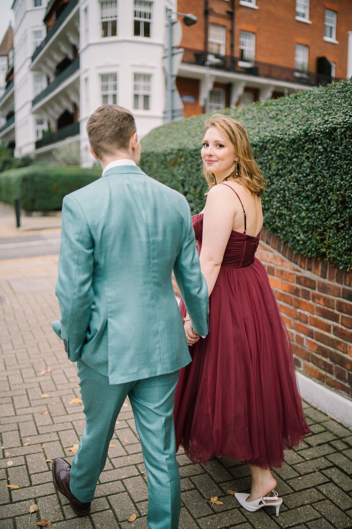 A couple in formal attire, a man in a teal suit and a woman in a burgundy dress, walking together on a brick path.