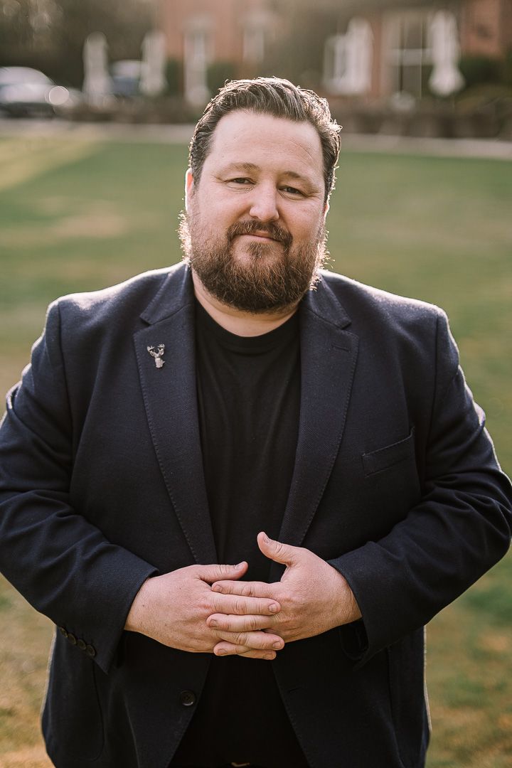 Bearded man in navy blazer and black shirt stands outdoors on green lawn, hands clasped, smiling confidently.