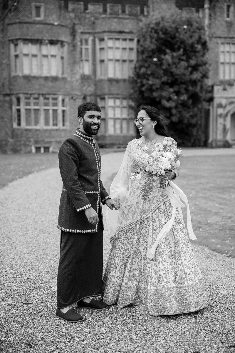 A couple in traditional wedding attire pose together on the grounds of a grand estate with a historic building in background.