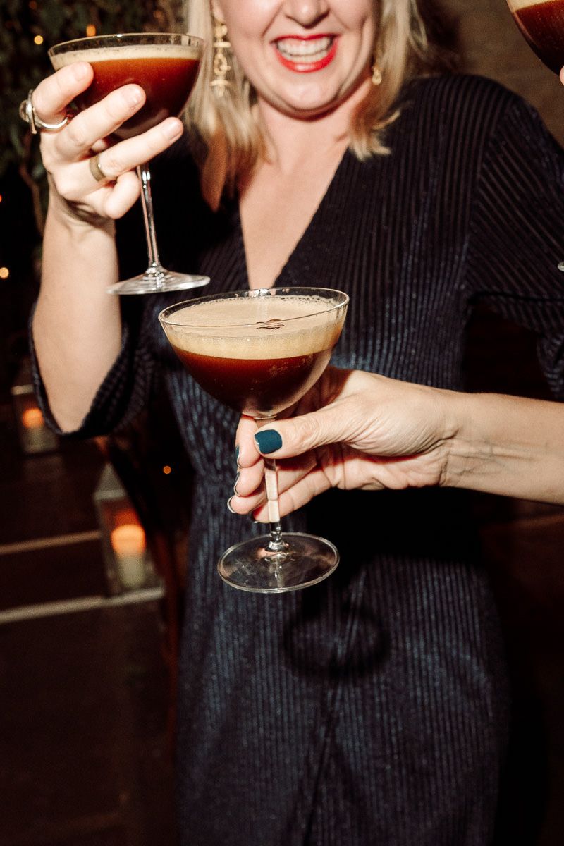 Someone in a black dress holds two cocktail glasses with espresso martinis at a dimly lit bar.