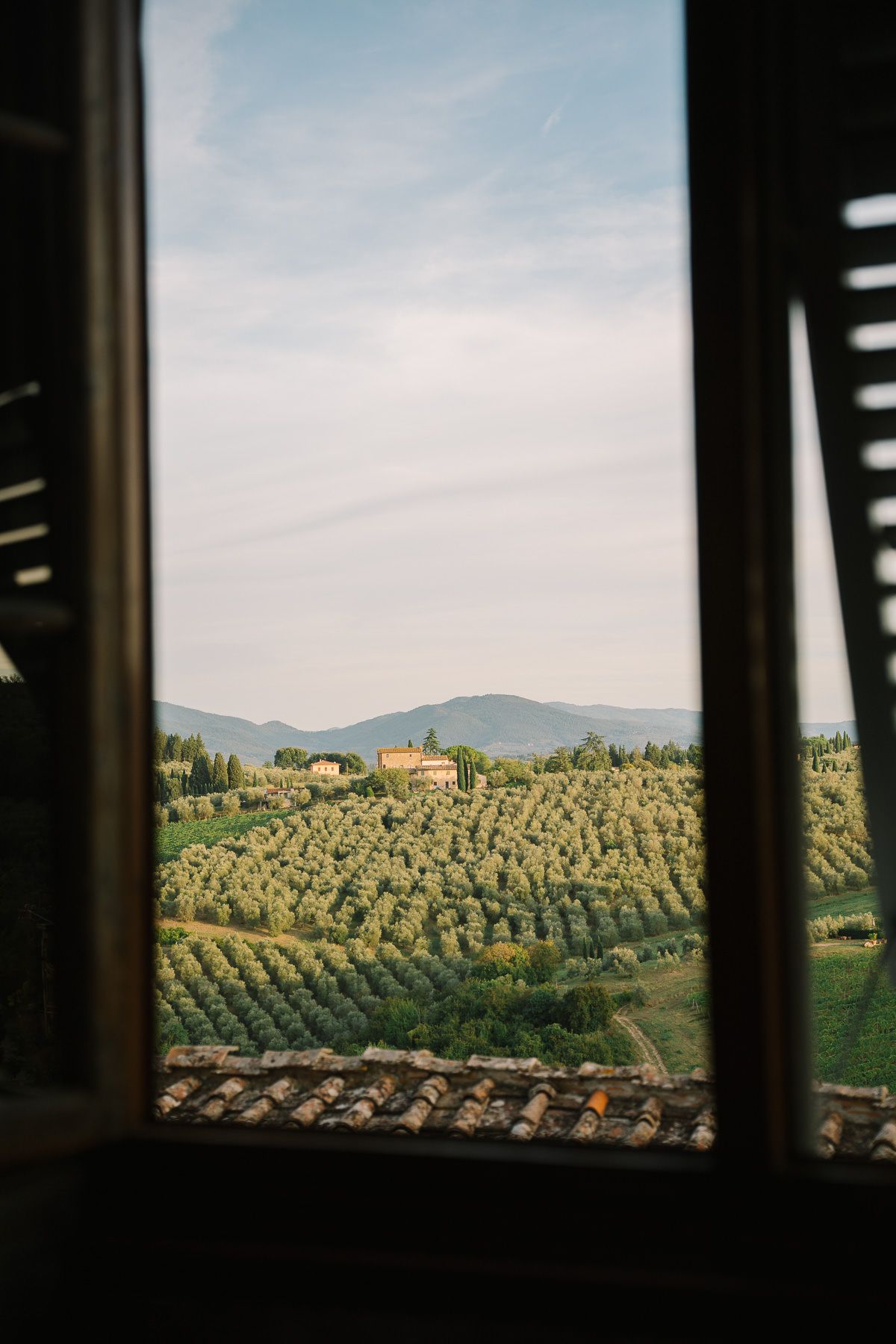 A scenic view of a rural Italian landscape with rolling hills, olive groves, and a distant village framed through a window.