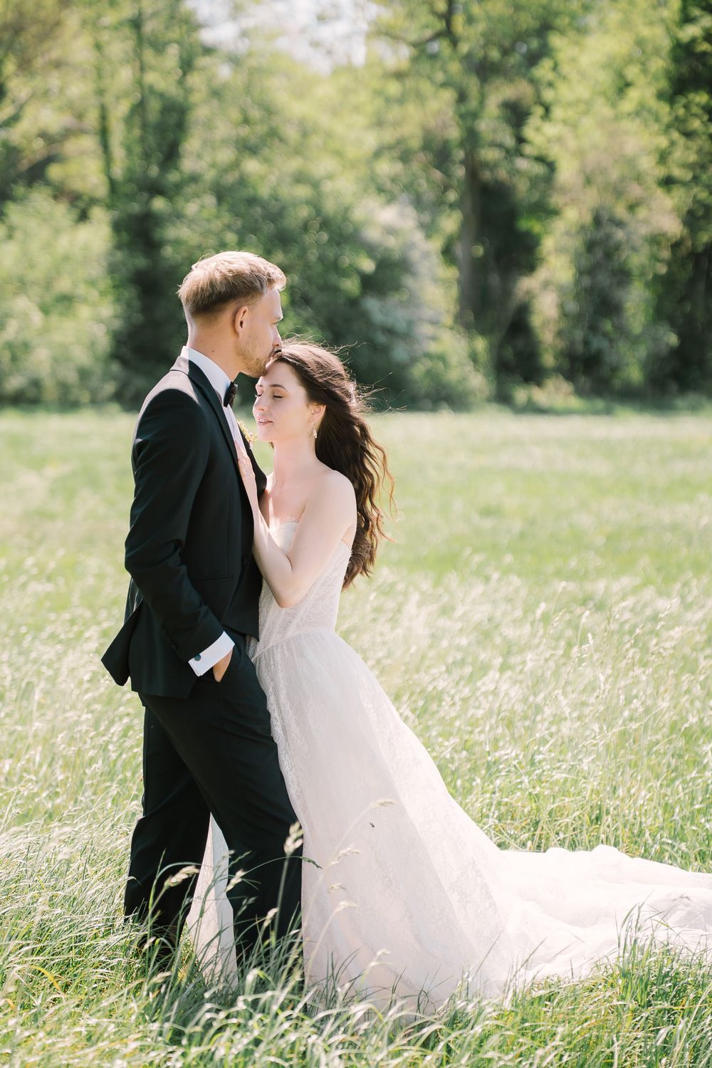 Bride in white gown and groom in black tuxedo embrace in a sunlit green meadow with lush trees in the background.