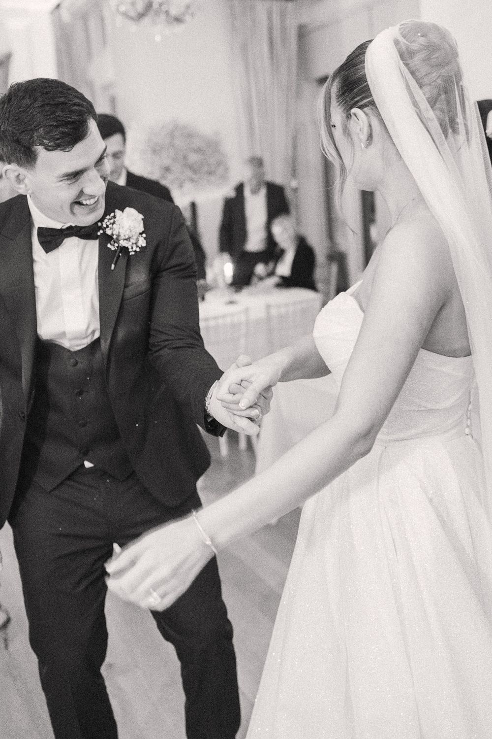Black and white photo of a smiling groom in a tuxedo dancing with his bride in a white gown and veil at their wedding reception.