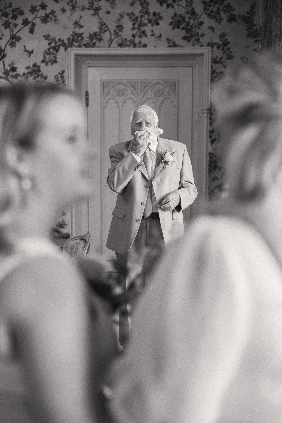 A black and white image of an elderly man in a suit standing in a room with ornate architectural details.