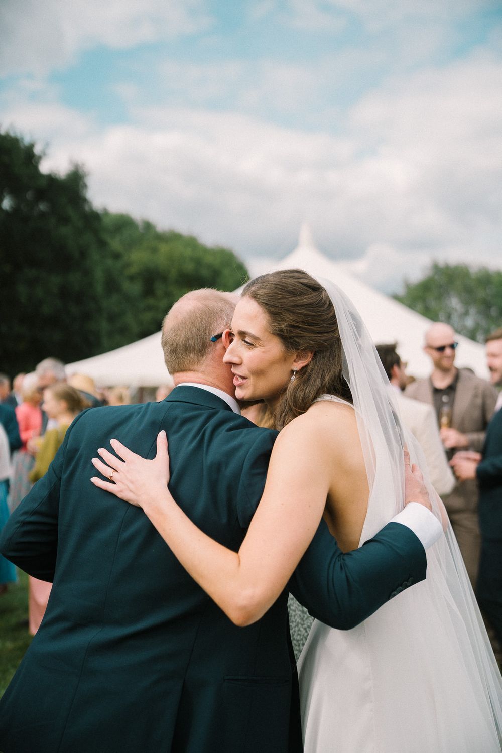 Bride in white gown and veil embracing a man in dark suit at outdoor wedding reception with tent.