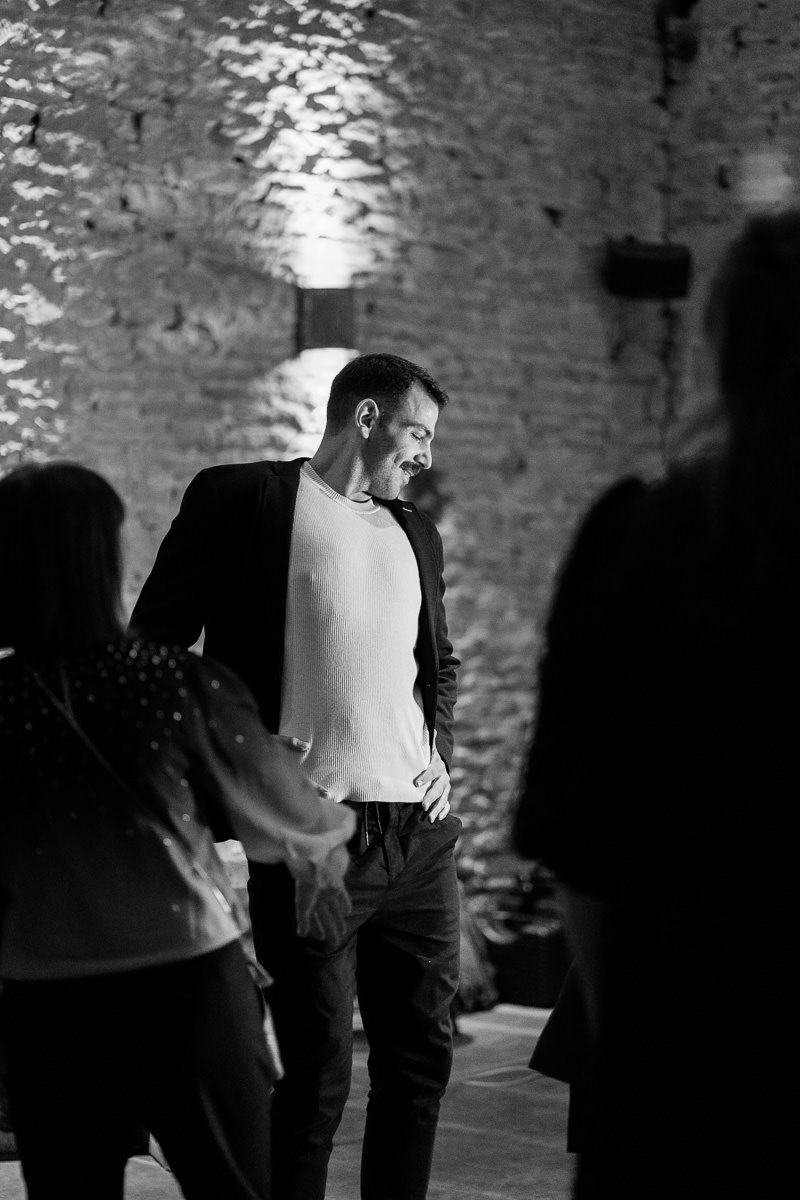 A performer in a black blazer and white shirt stands on stage against a textured brick wall in dramatic lighting.