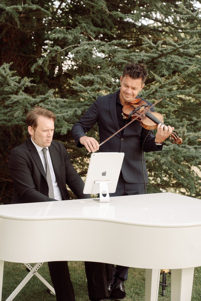 Two musicians perform at an outdoor event with one playing violin and another at a white piano against an evergreen backdrop.