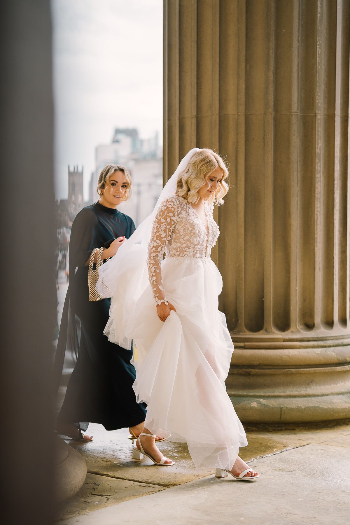 A woman in a white wedding dress and a man in a black suit walking together near a stone column.