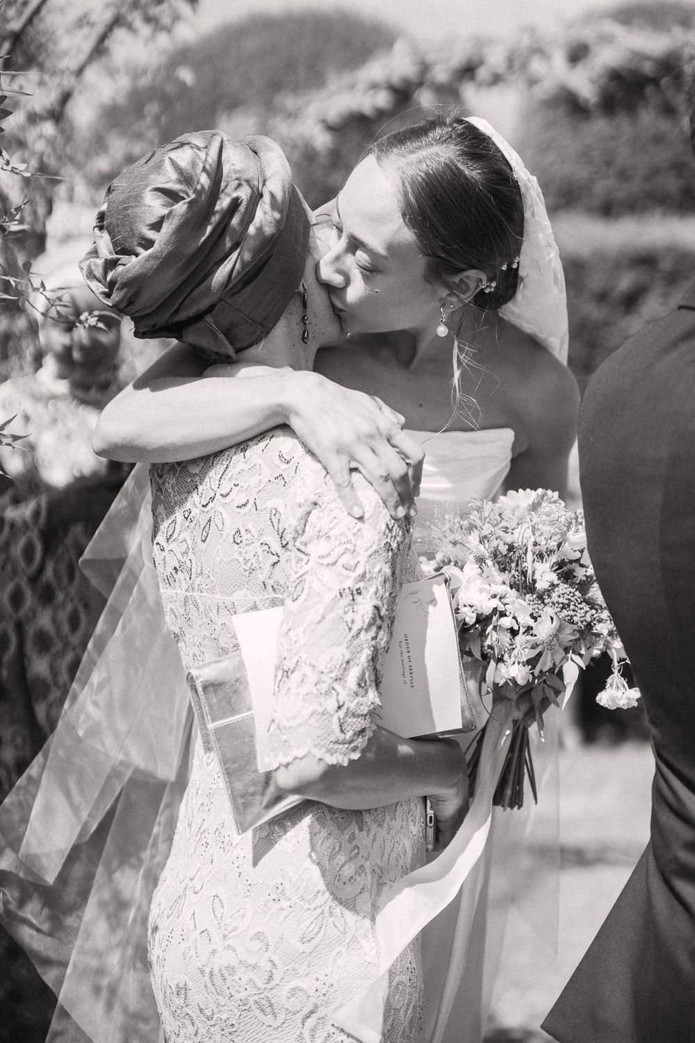 Black and white photo of two women embracing and kissing at a wedding, one in a lace gown holding a bouquet.