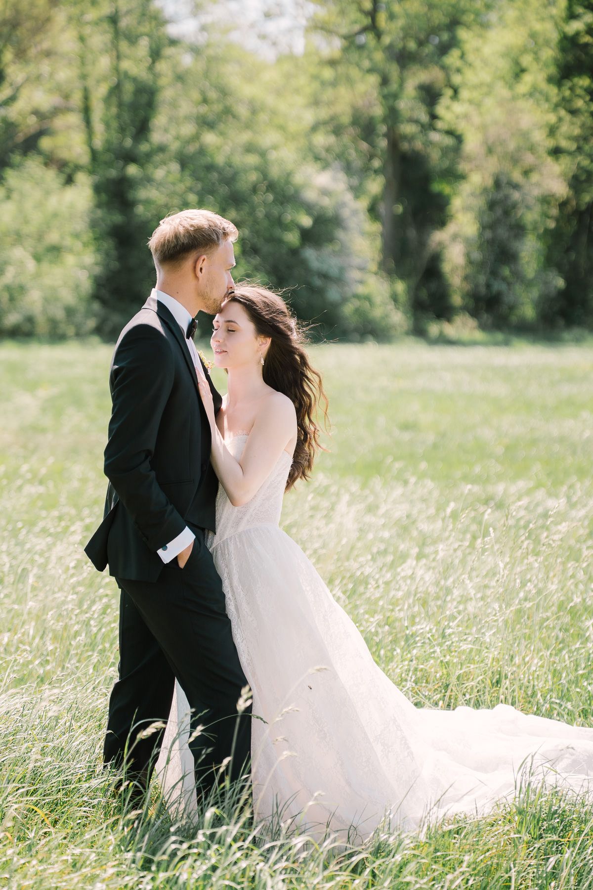 A bride and groom embrace in a grassy field, surrounded by trees and nature.
