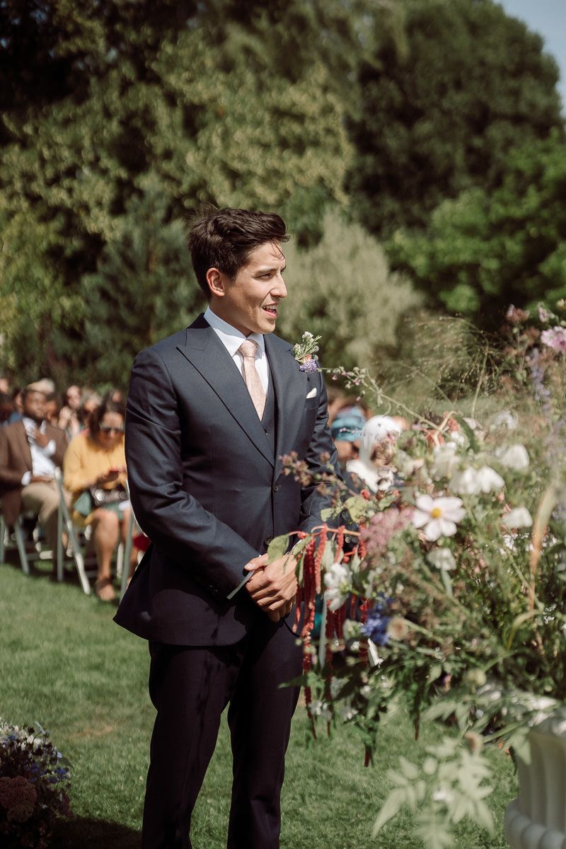 A groom in a dark suit and pink tie stands at the altar during an outdoor garden wedding ceremony.