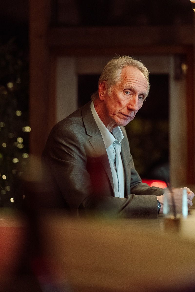 A person in a gray suit and light blue shirt sits at a table in a dimly lit room.