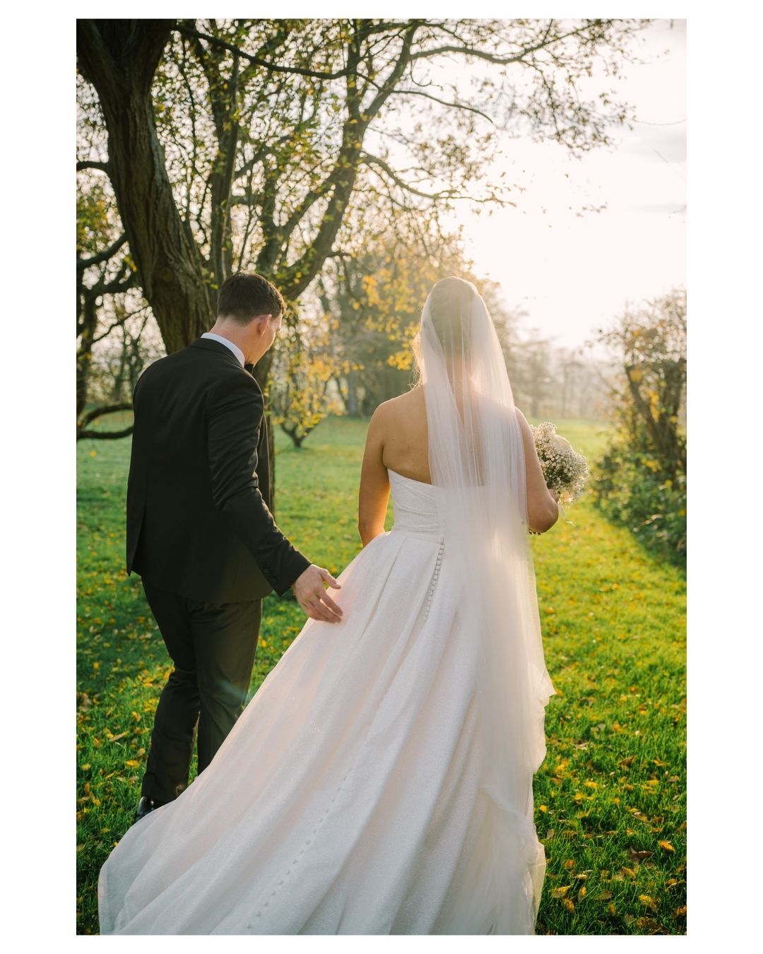 A bride and groom walk hand-in-hand through a sunlit garden, she in a flowing white gown with veil.