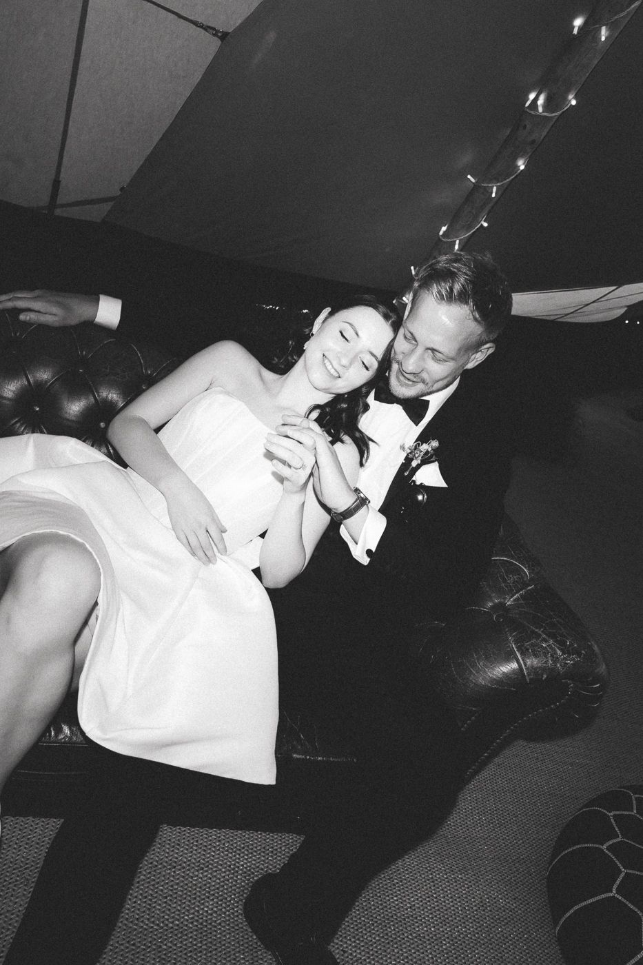 A couple in formal wedding attire relaxes together on a dark leather couch at their reception.