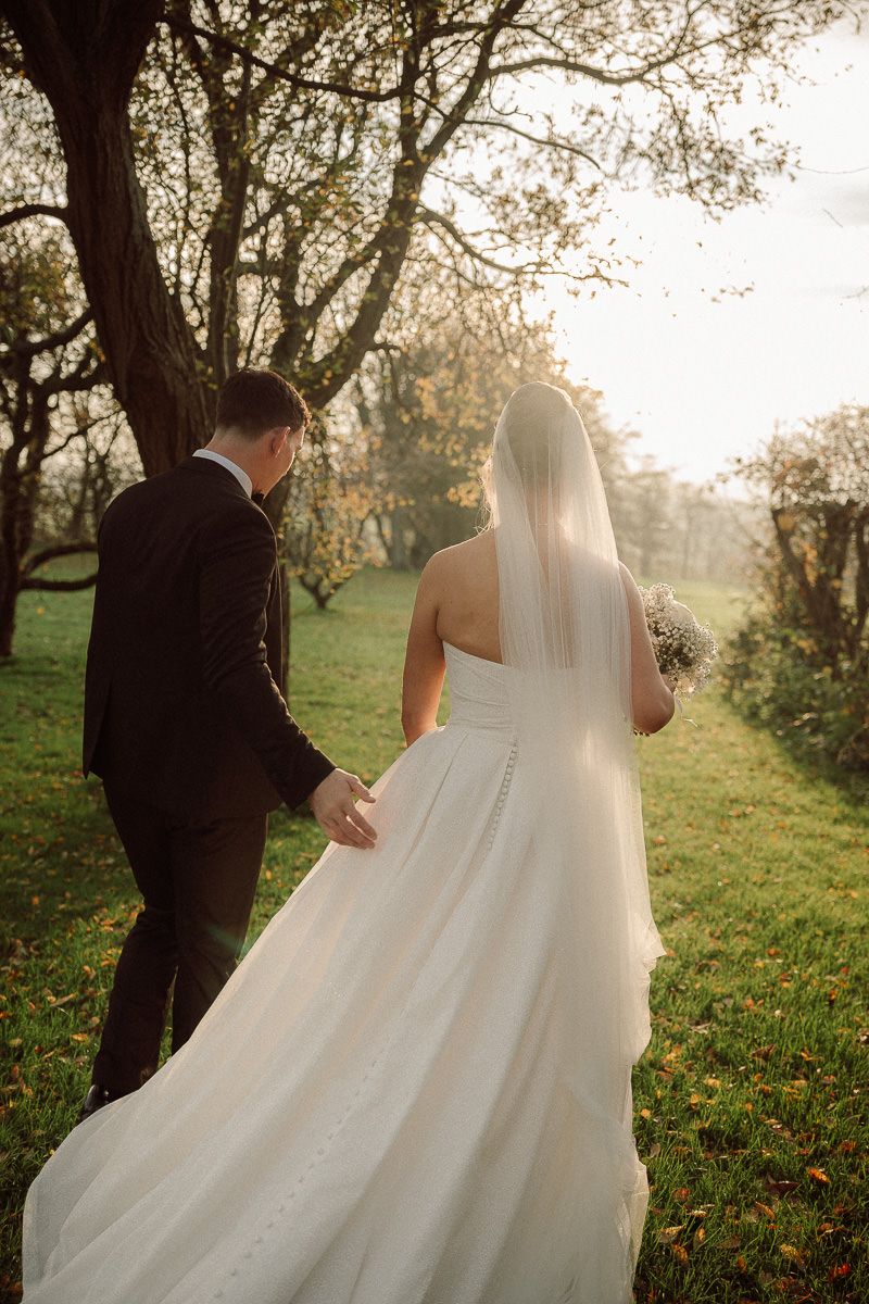 A couple in wedding attire walks through a sunlit autumn meadow with trees in the background.