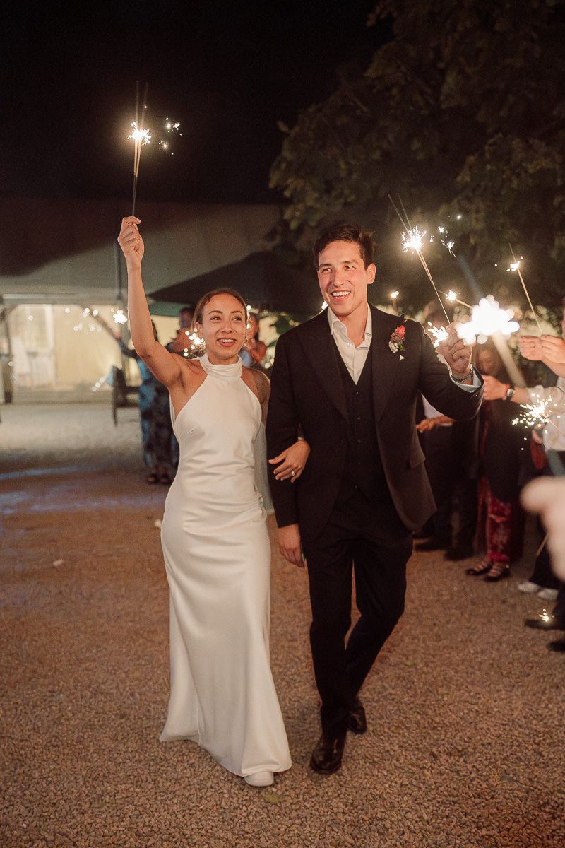 Wedding couple celebrates with sparklers during their nighttime reception on gravel path.
