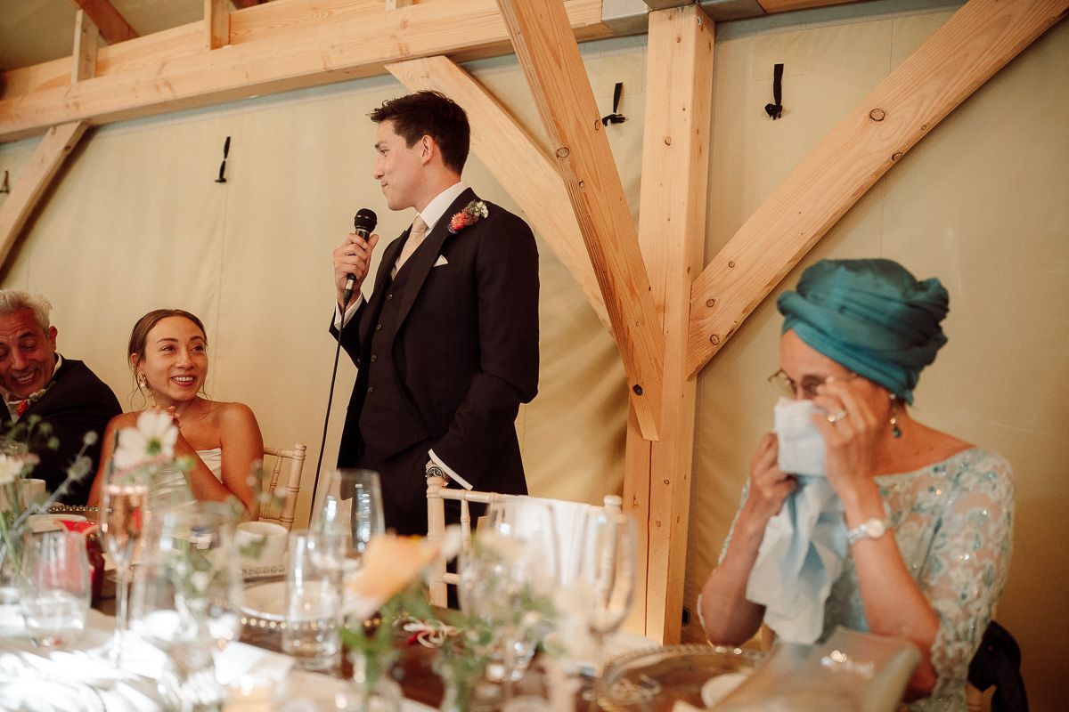 A wedding reception speech being given at a rustic wooden-beamed venue with decorated dining tables.