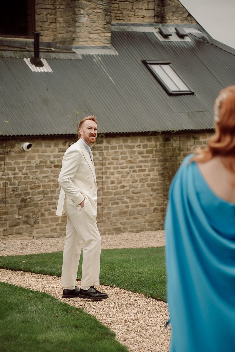 A person in a white suit stands on a gravel path next to a stone wall and grassy lawn.