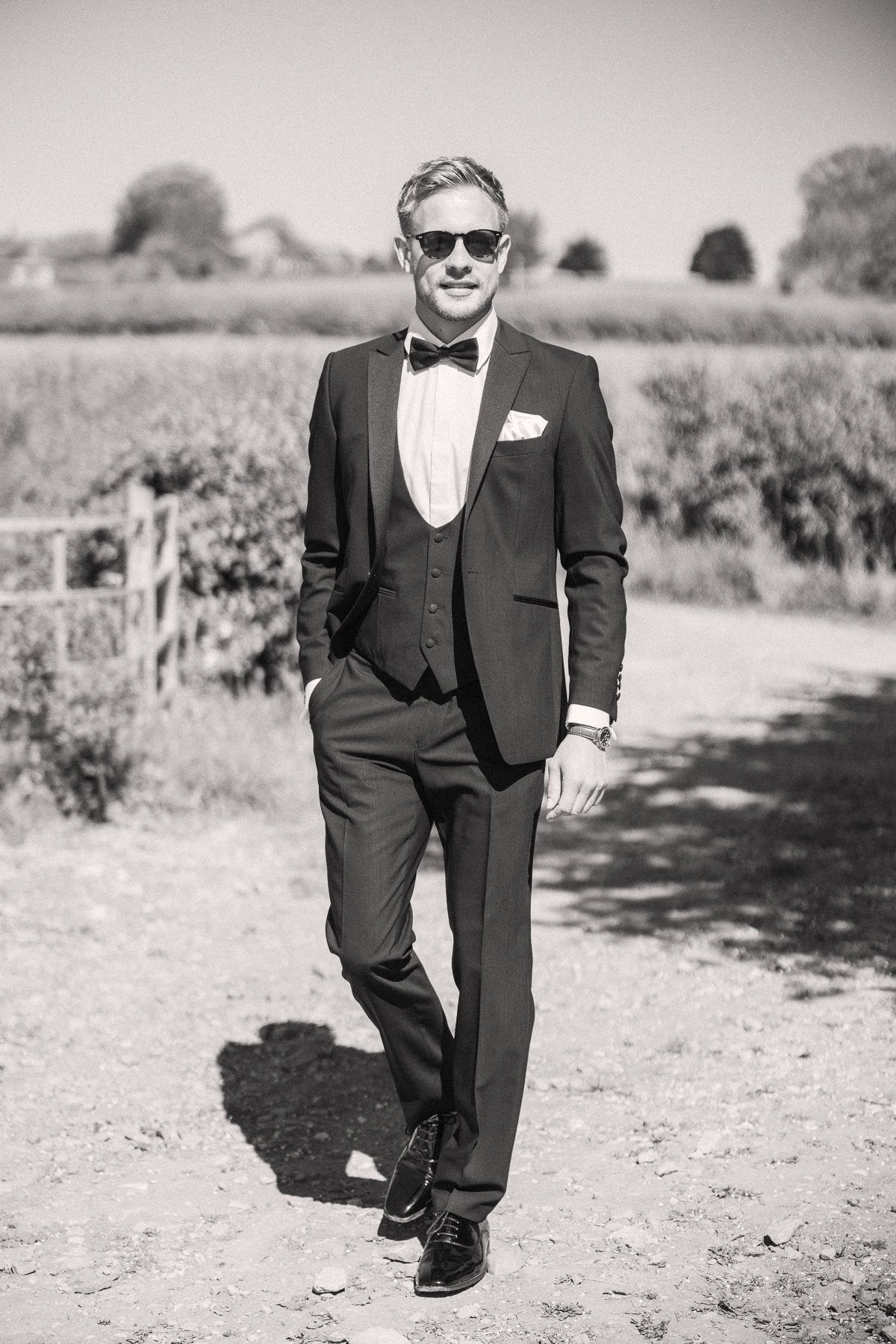 A man in a tuxedo and bowtie stands in a field, looking confident and stylish.