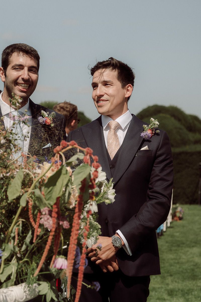 Two people in dark suits share joyful moments outdoors with floral arrangements at a wedding celebration.