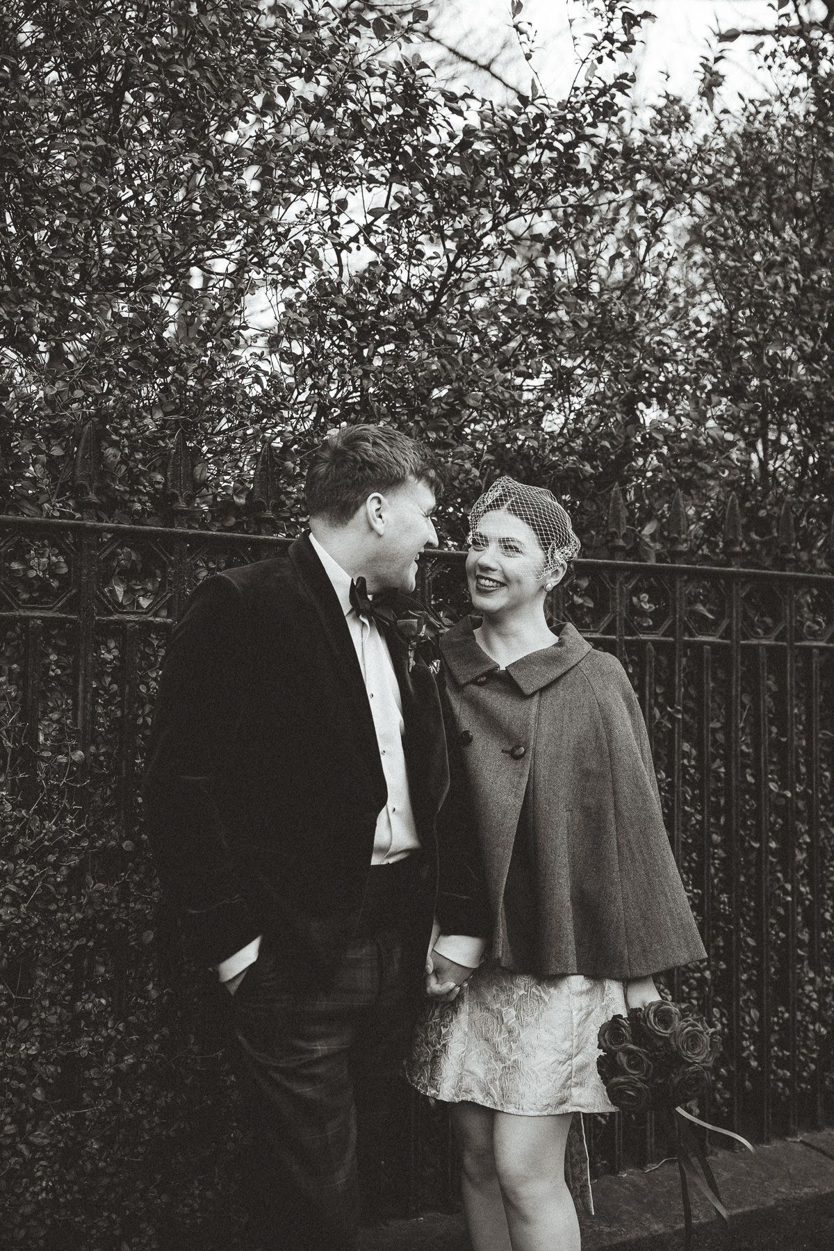 Two figures in vintage attire stand together in a garden against an iron fence, sharing a candid black and white moment.