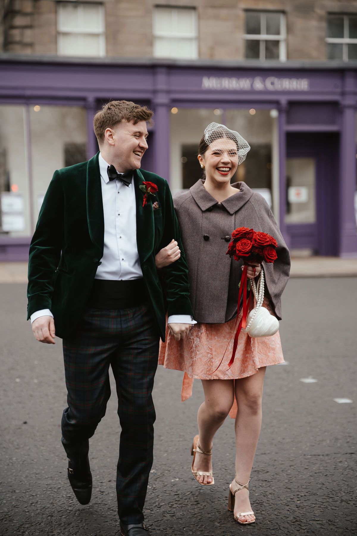 A couple laughs while walking down a city street, with a red bouquet and formal attire on a rainy day.