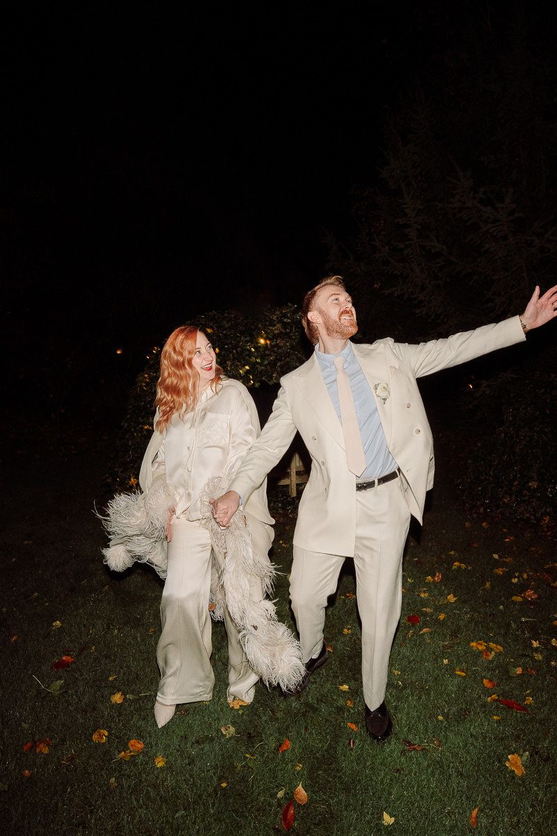A couple in white attire dances playfully on a grass lawn with autumn leaves scattered around at night.