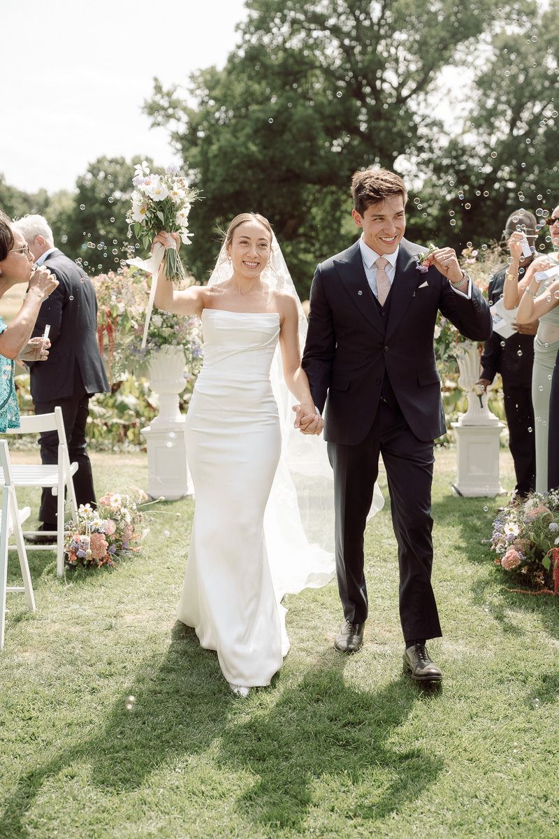 Newlyweds walk down aisle together on grassy lawn while guests celebrate with flower petals.