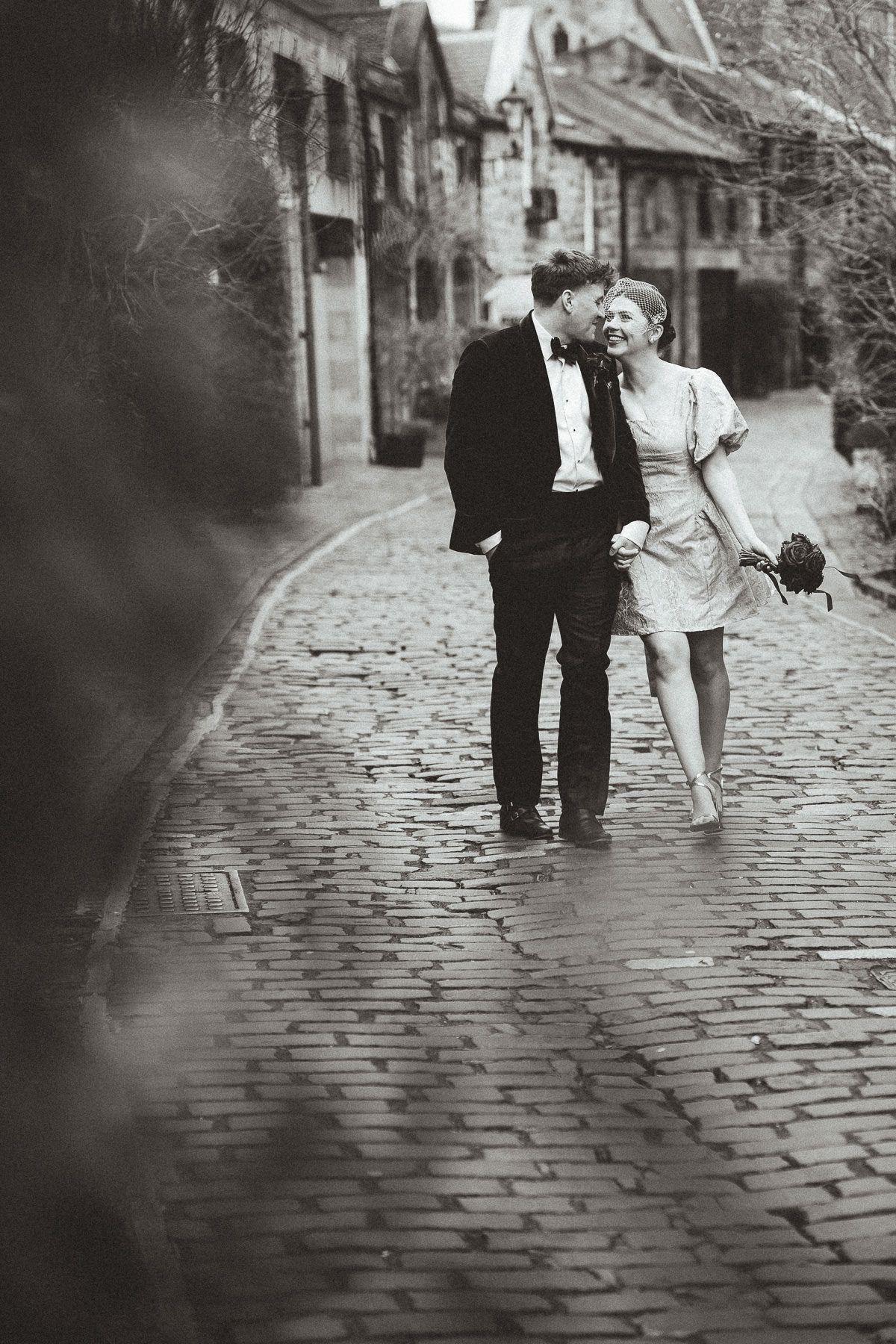A couple walks together on a cobblestone street in black and white, creating a romantic vintage atmosphere.