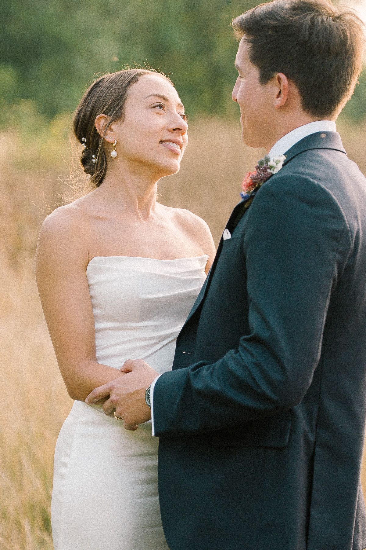 A bride in a strapless white gown smiles at her groom in a navy suit, holding hands in a golden field.
