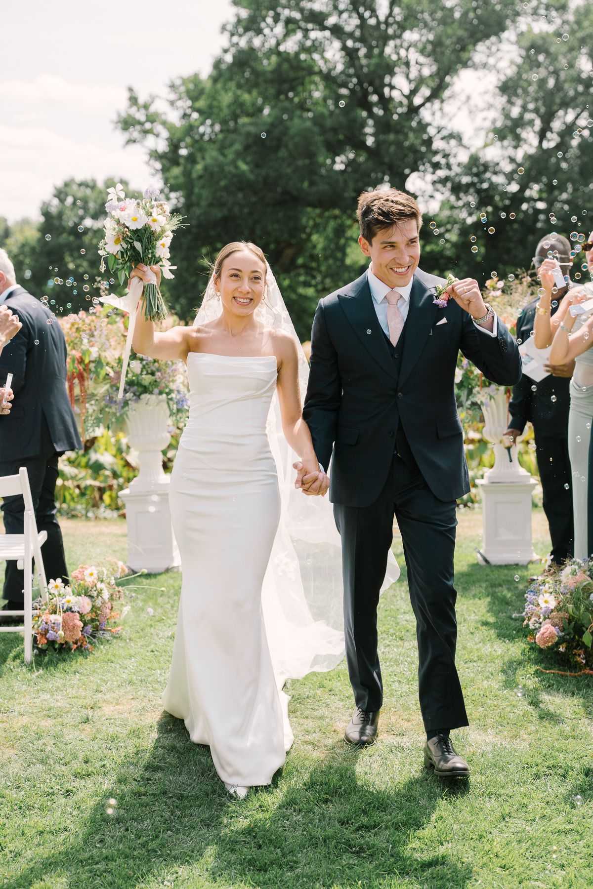 A bride and groom walking together on a grassy lawn surrounded by wedding guests.