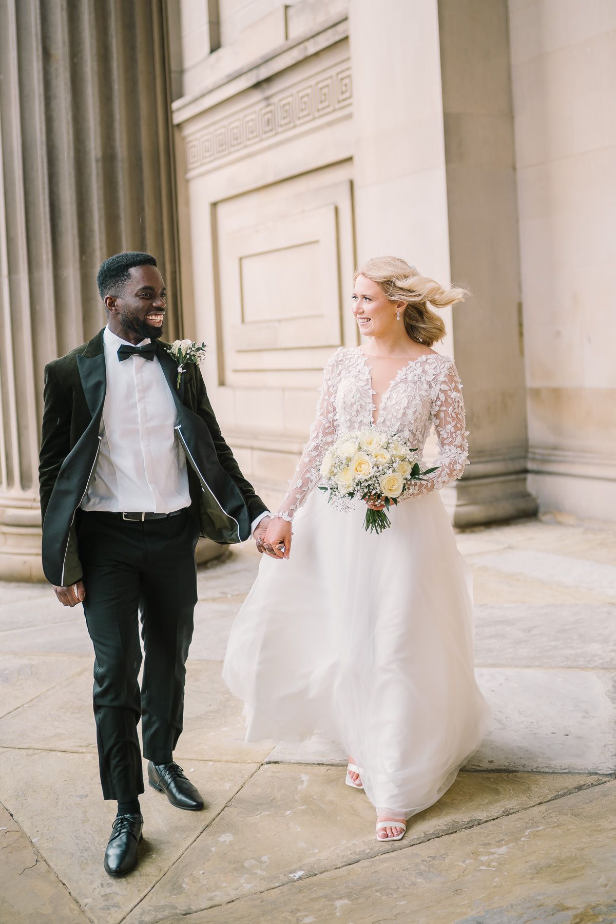 A bride and groom walk together on the steps of a building, the bride wearing a lace wedding dress and the groom wearing a black tuxedo.