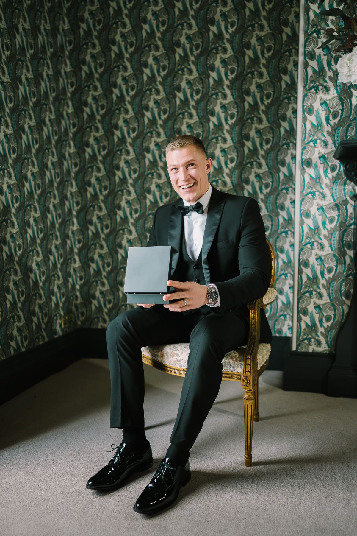 A well-dressed man in a tuxedo sits in a chair, holding a laptop and smiling at the camera.