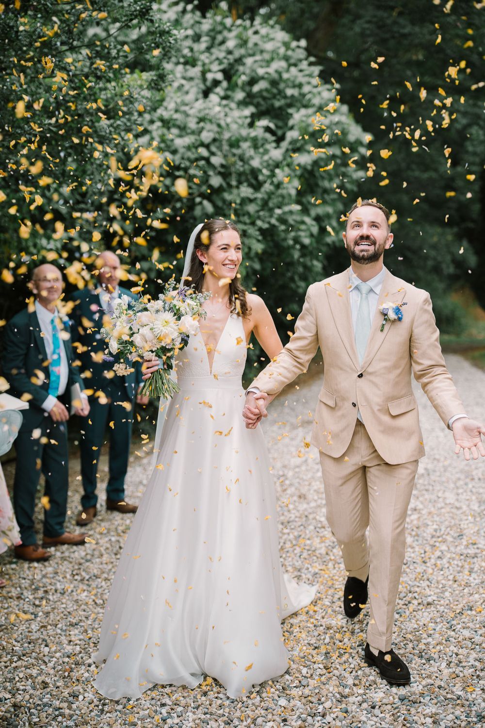 Bride and groom walk hand-in-hand through yellow confetti shower on gravel path, guests celebrating in background.