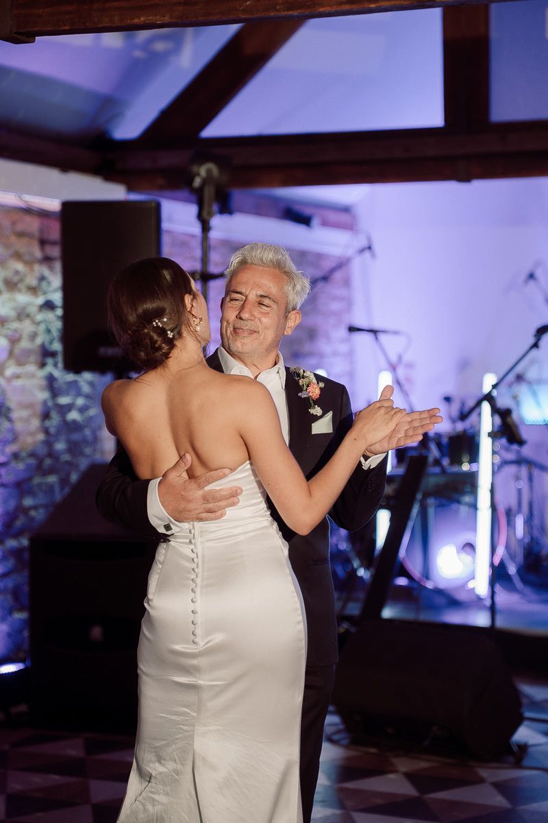 A couple shares an intimate dance at their wedding reception, with the bride wearing a white dress with button details.