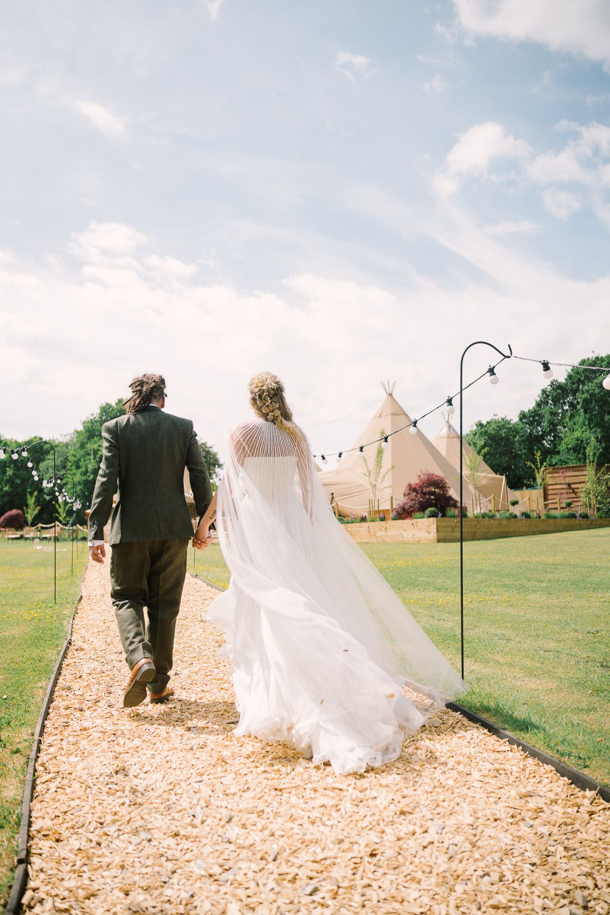 A bride and groom walk down a gravel path in a grassy field, with a teepee structure in the background.