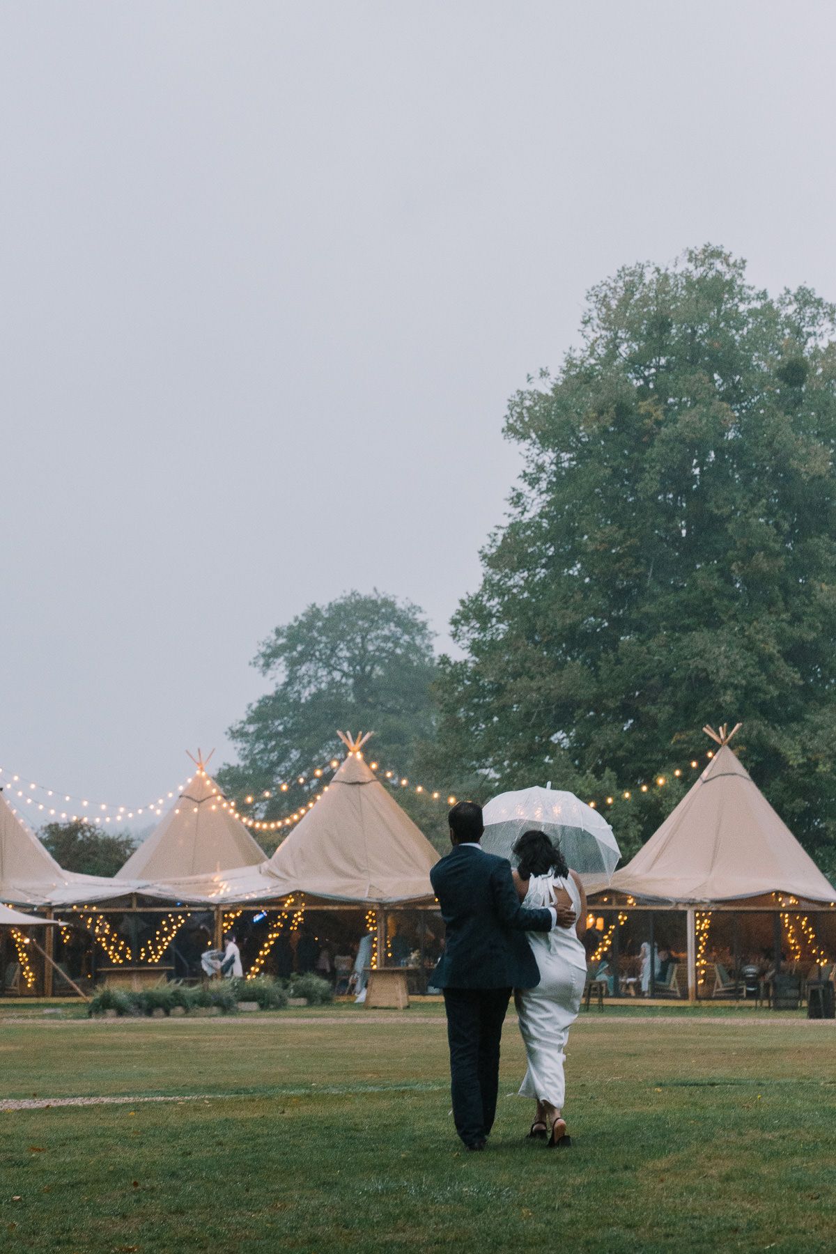 A wedding ceremony with white tents and string lights in a grassy field surrounded by trees.