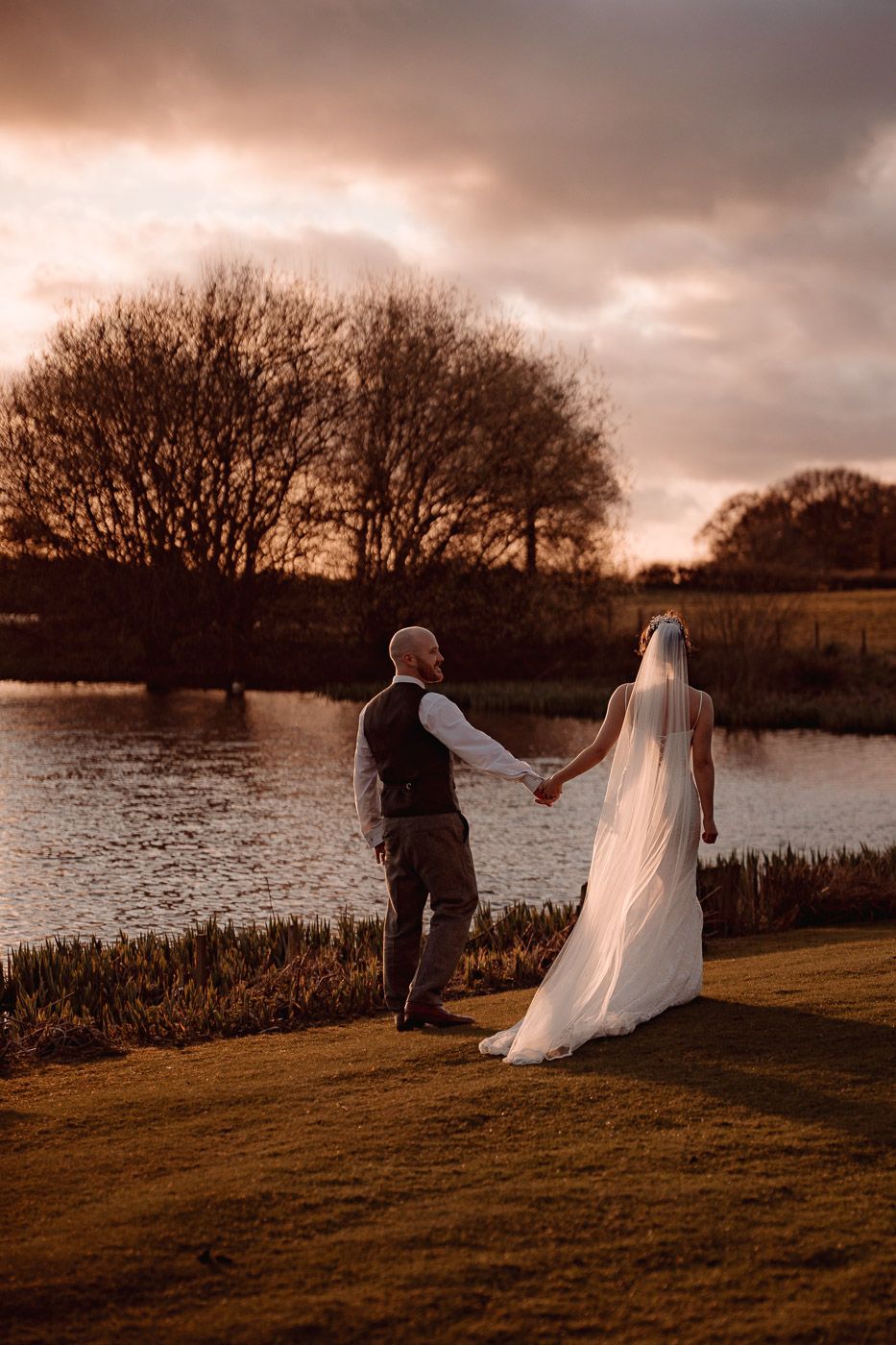 A romantic sunset wedding photo showing a couple holding hands by a serene lake with dramatic orange sky.