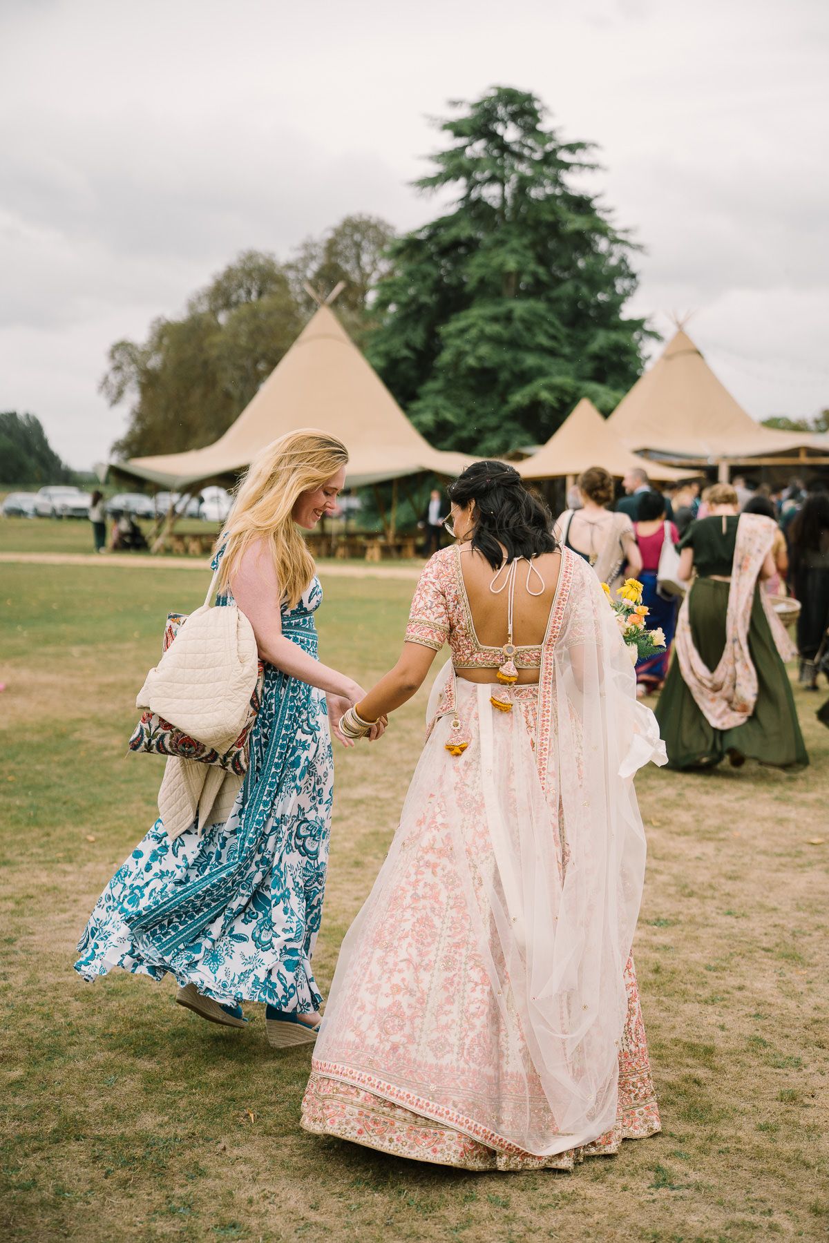 Two women in colorful traditional Indian dresses are walking together on a grassy field with tents in the background.