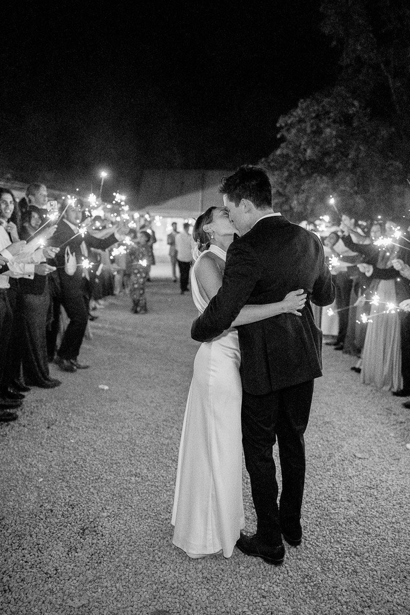 A couple in wedding attire dances together at night while surrounded by sparklers held by wedding guests.