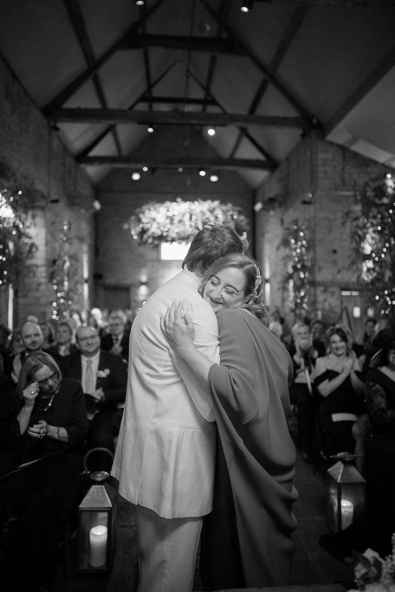 A tender embrace between two people during a wedding ceremony in a rustic venue with exposed beams and twinkling lights.